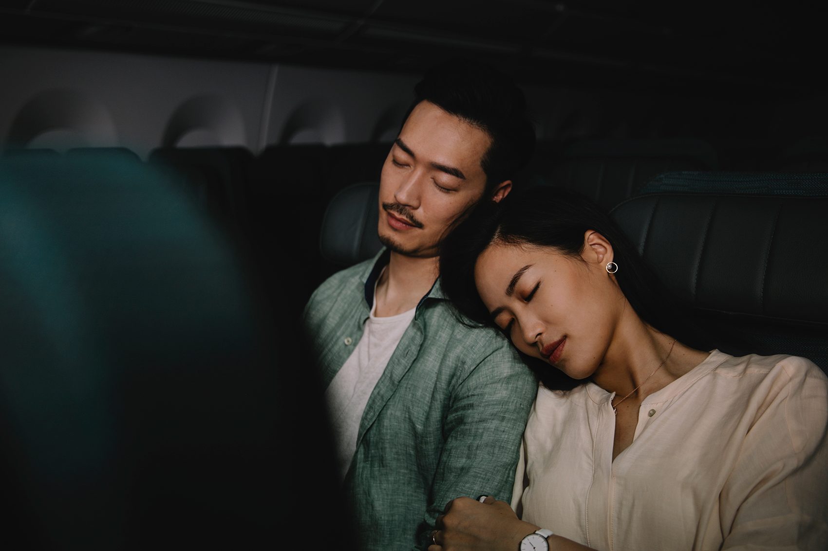 A man and a woman sleeping on a plane. The woman’s head is on the man’s shoulder. 