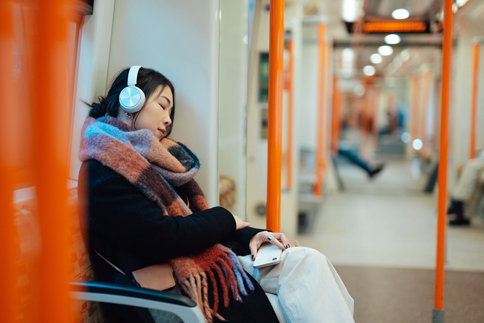 A woman wearing headphones and a plaid scarf, sleeping on a train while holding her phone. 