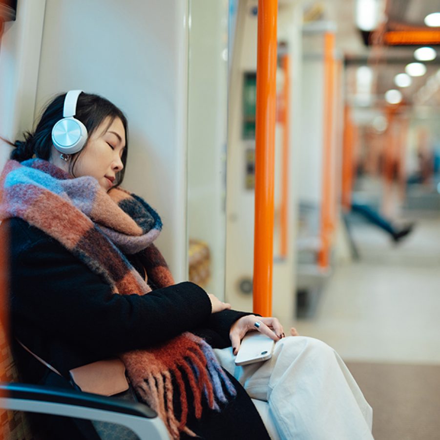 A woman wearing headphones and a plaid scarf, sleeping on a train while holding her phone. 