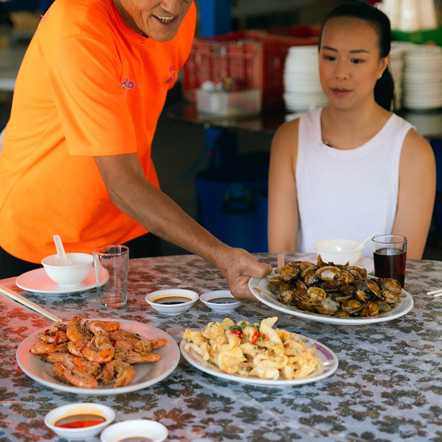 A man serves a plate of clams to a woman at Ming Kee Seafood Restaurant on Po Toi island. On the table are plates of prawns and fried squid. 