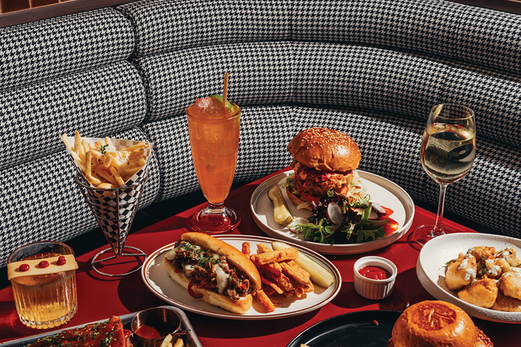 A red table at American diner RightSideUp filled with food and drinks like burgers, hot dogs, and fries in front of a houndstooth booth