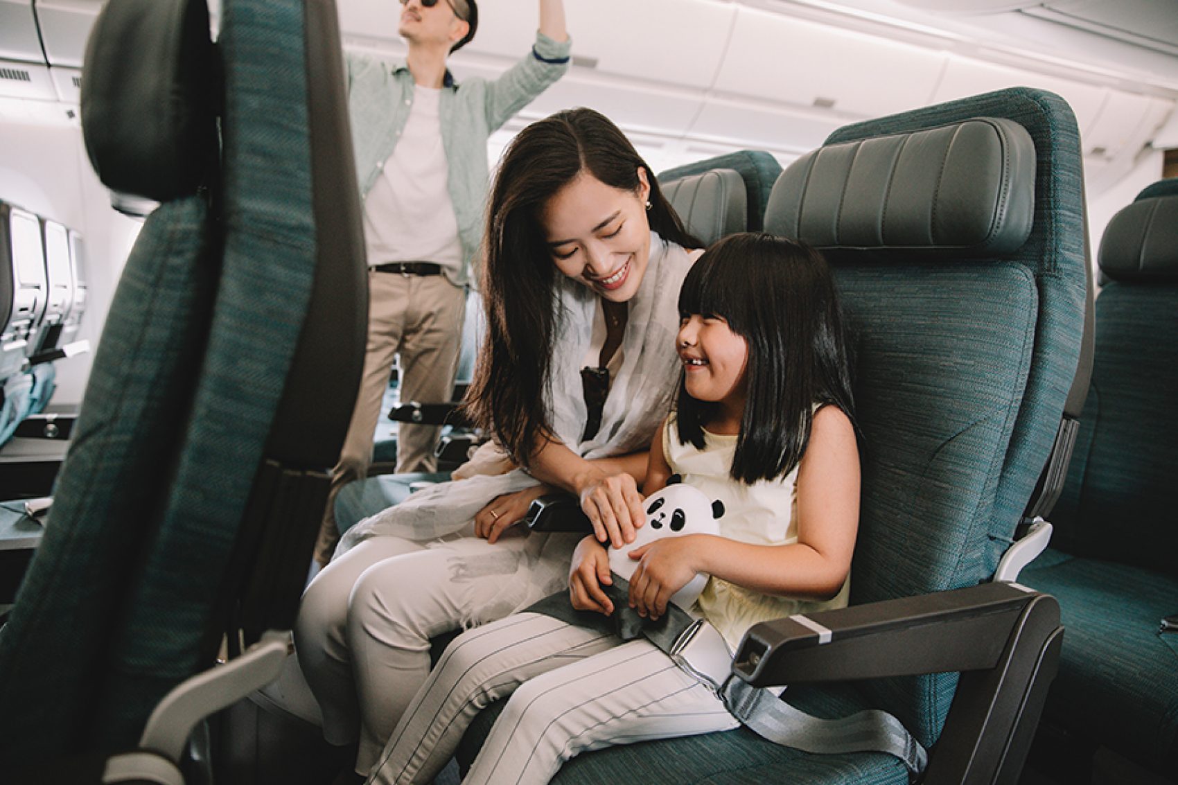 A family is on a Cathay Pacific flight, with the mom and daughter seated and holding a MIN MIN panda toy while the father is storing luggage in the overhead bin
