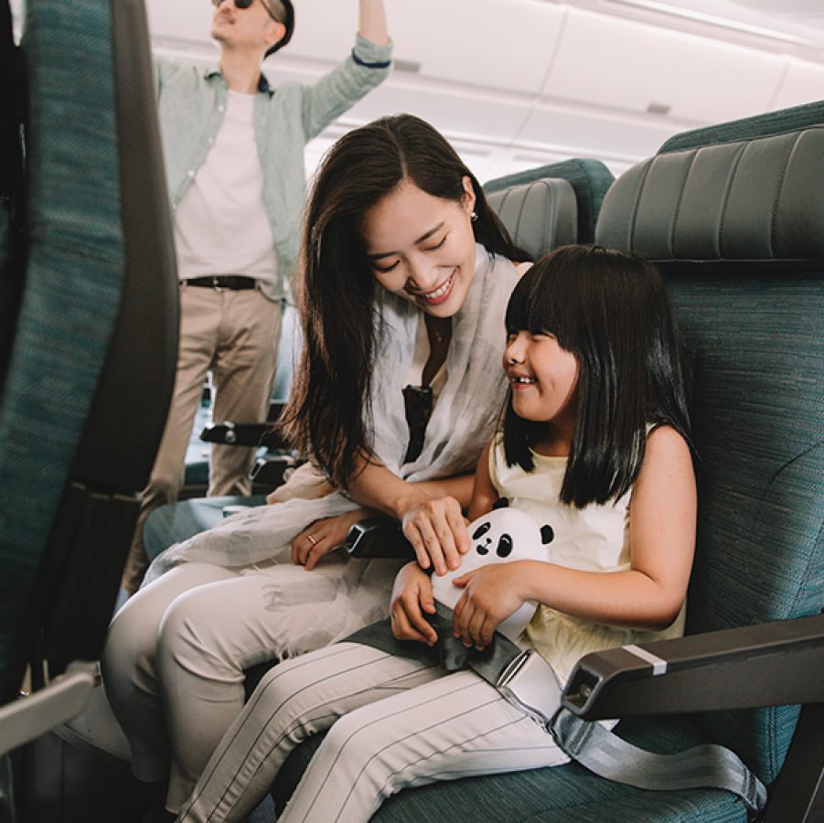 A family is on a Cathay Pacific flight, with the mom and daughter seated and holding a MIN MIN panda toy while the father is storing luggage in the overhead bin