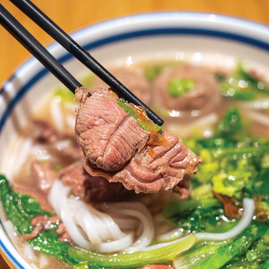  A close-up of a pair of chopsticks holding a piece of sliced beef over a bowl of soup noodles