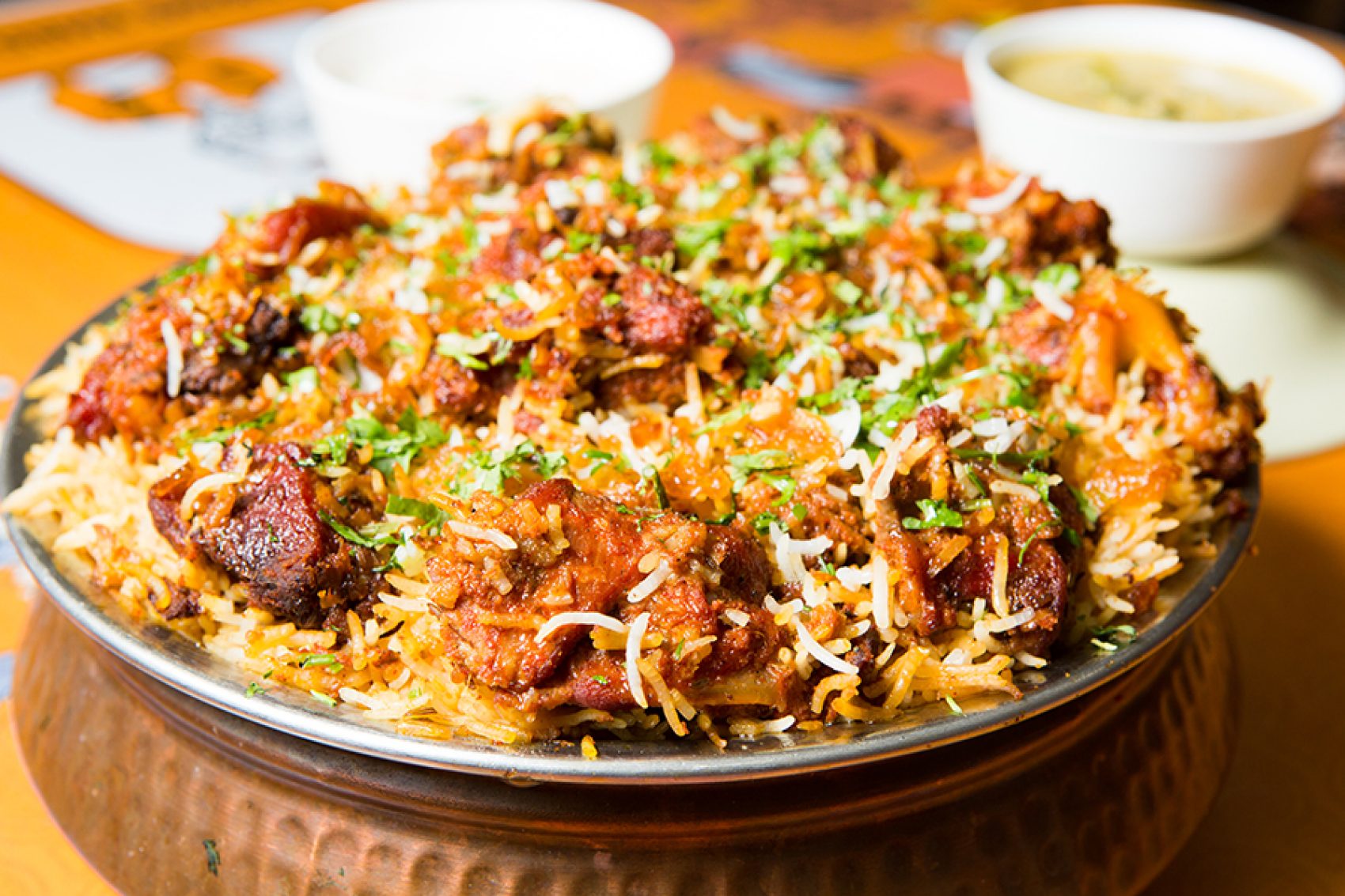 A close up of Hyderabadi meat biryani served in a silver bowl for sharing on a dining table along side two bowls of soup    