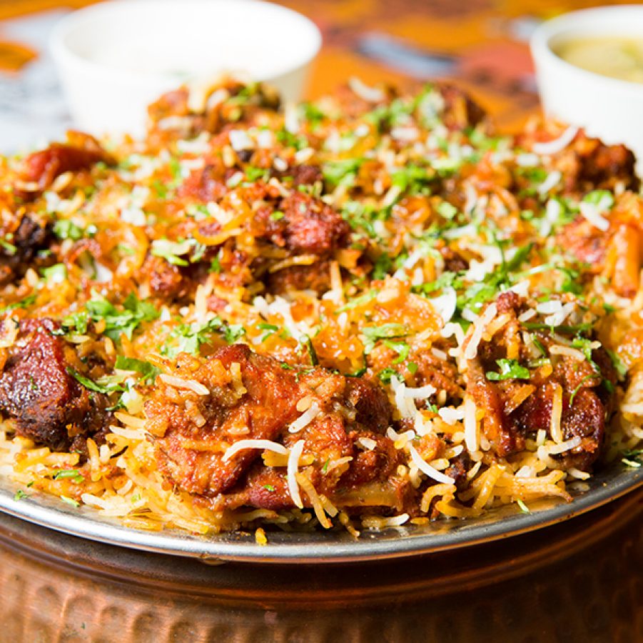 A close up of Hyderabadi meat biryani served in a silver bowl for sharing on a dining table along side two bowls of soup    