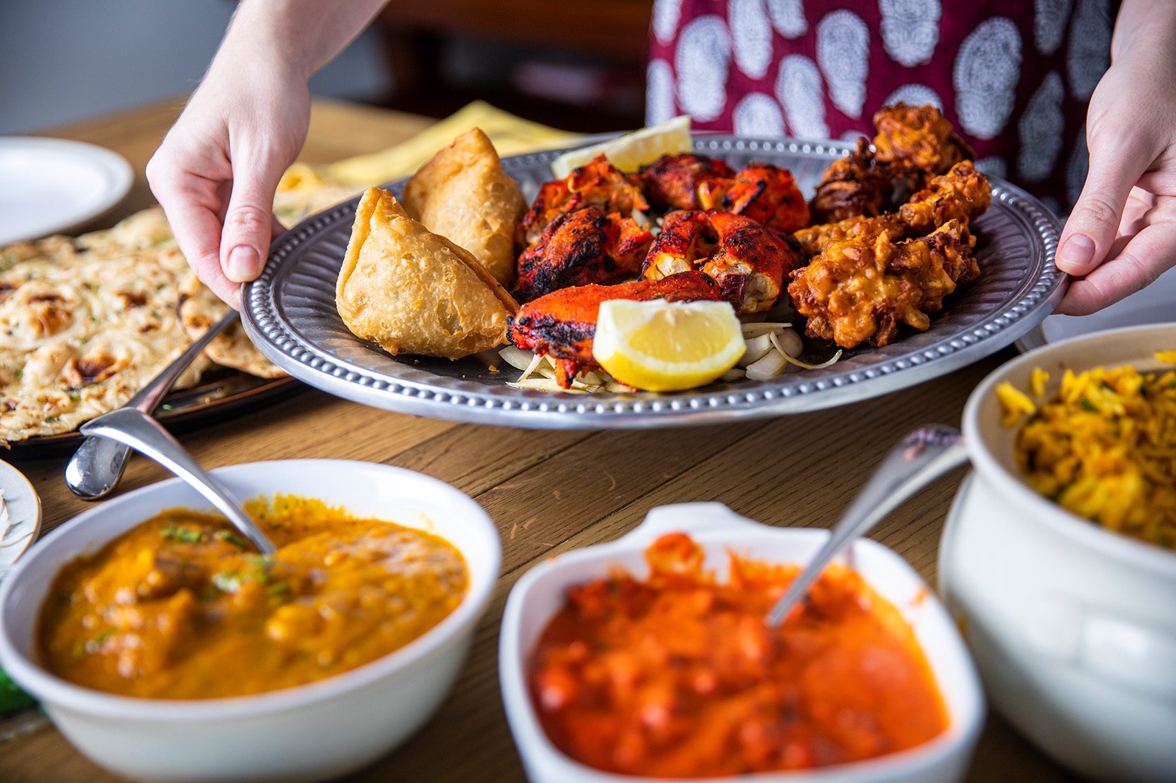 A dining table full of Indian dishes; curry, pratha, samosa, tandoori chicken, pakora on a silver platter