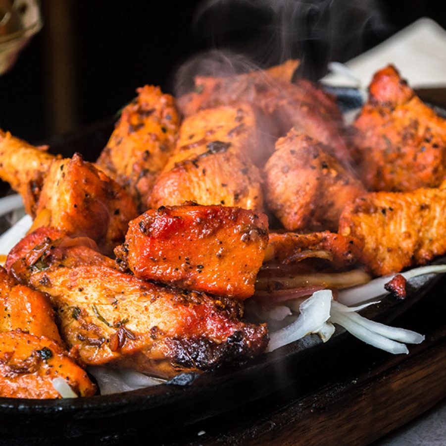 A platter of sizzling tandoori chicken served ontop of onions on a black iron plate    