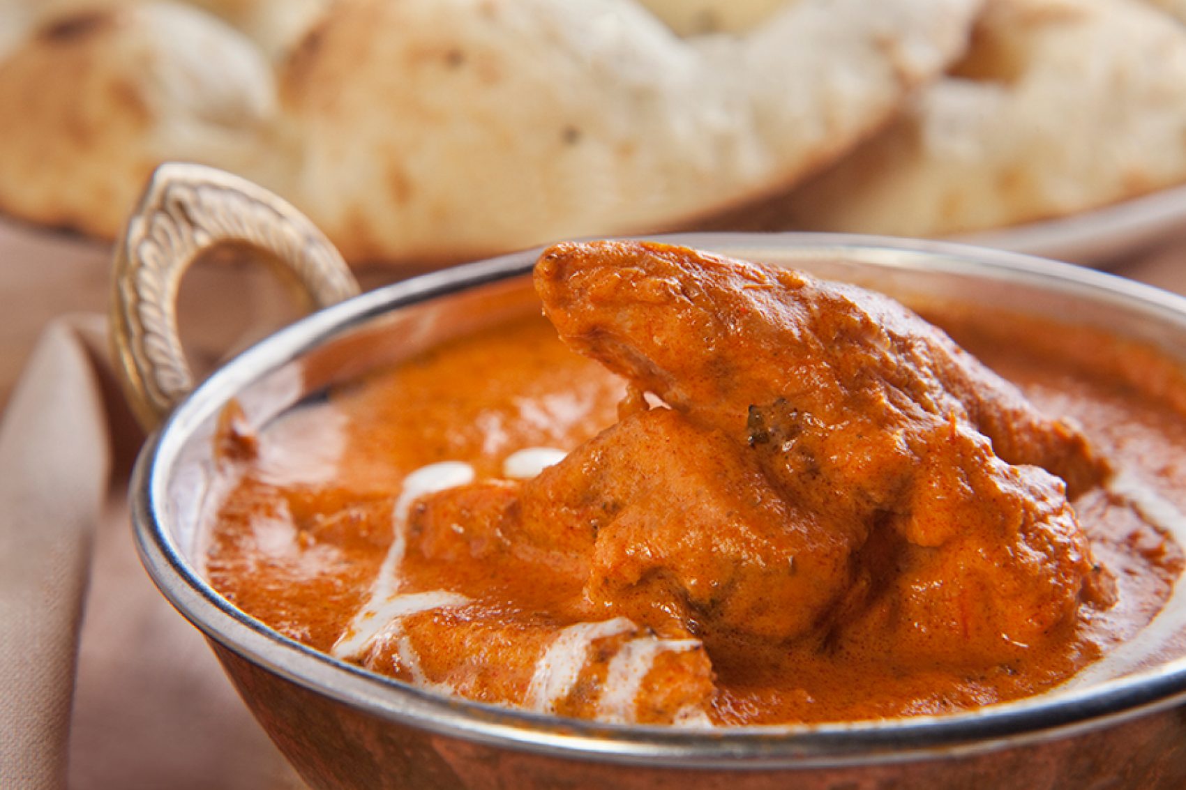 A closeup of butter chicken in a silver bowl alongside a plat of naan 
