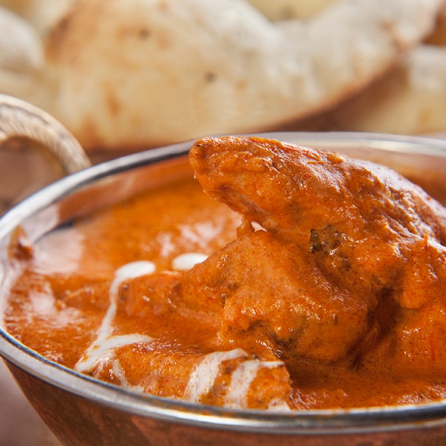 A closeup of butter chicken in a silver bowl alongside a plat of naan 