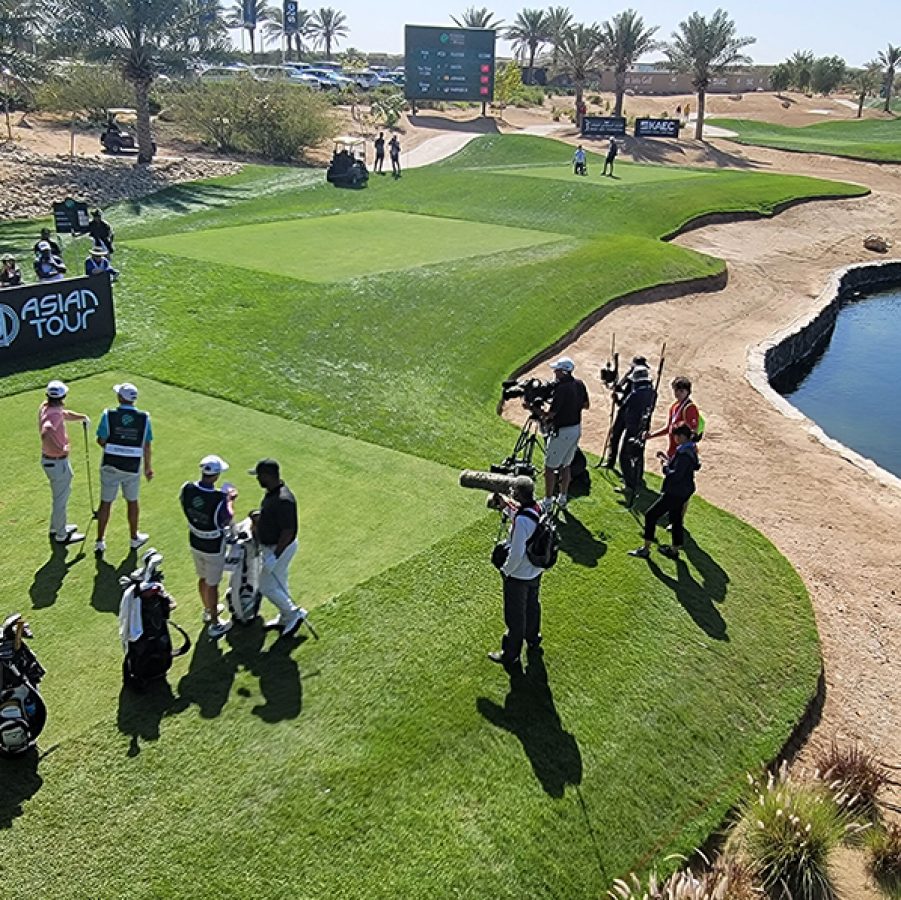 Golfers standing on grass and being filmed by a crew while spectators look on at Safaa Golf Club