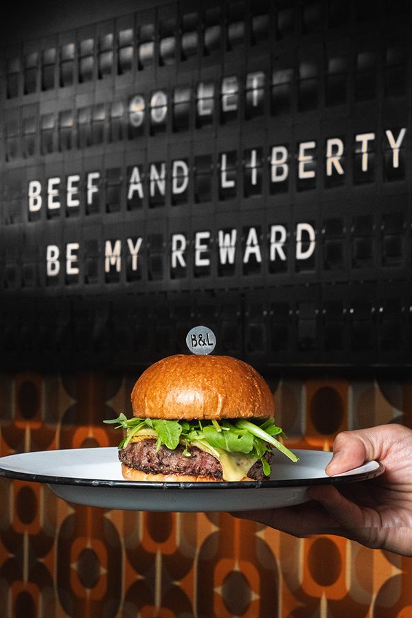 A hand holds a plate with a hamburger. The board in the background has white text spelling out “BEEF AND LIBERTY BE MY REWARD”.    