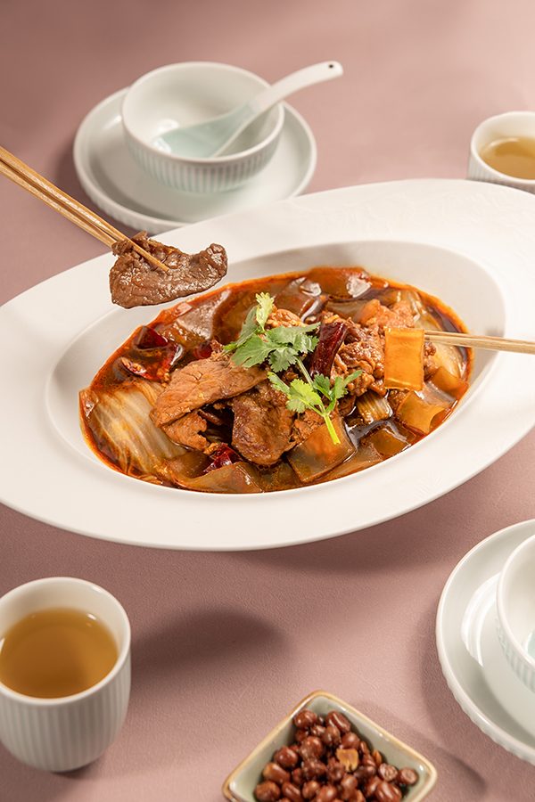 Chopsticks pick up morsels from a plate of poached beef in spicy sauce at Crystal Jade, Hong Kong. Two empty bowls with spoons, teacups and a small dish of peanuts surround the main dish. 