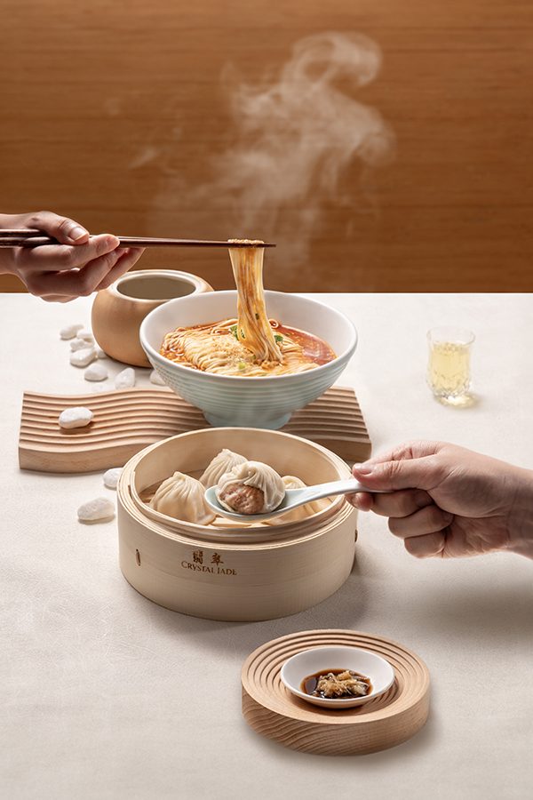 A bowl of dan dan noodles and a basket of xiao long bao at Crystal Jade, Hong Kong. Hands are picking up the food with chopsticks and a spoon.