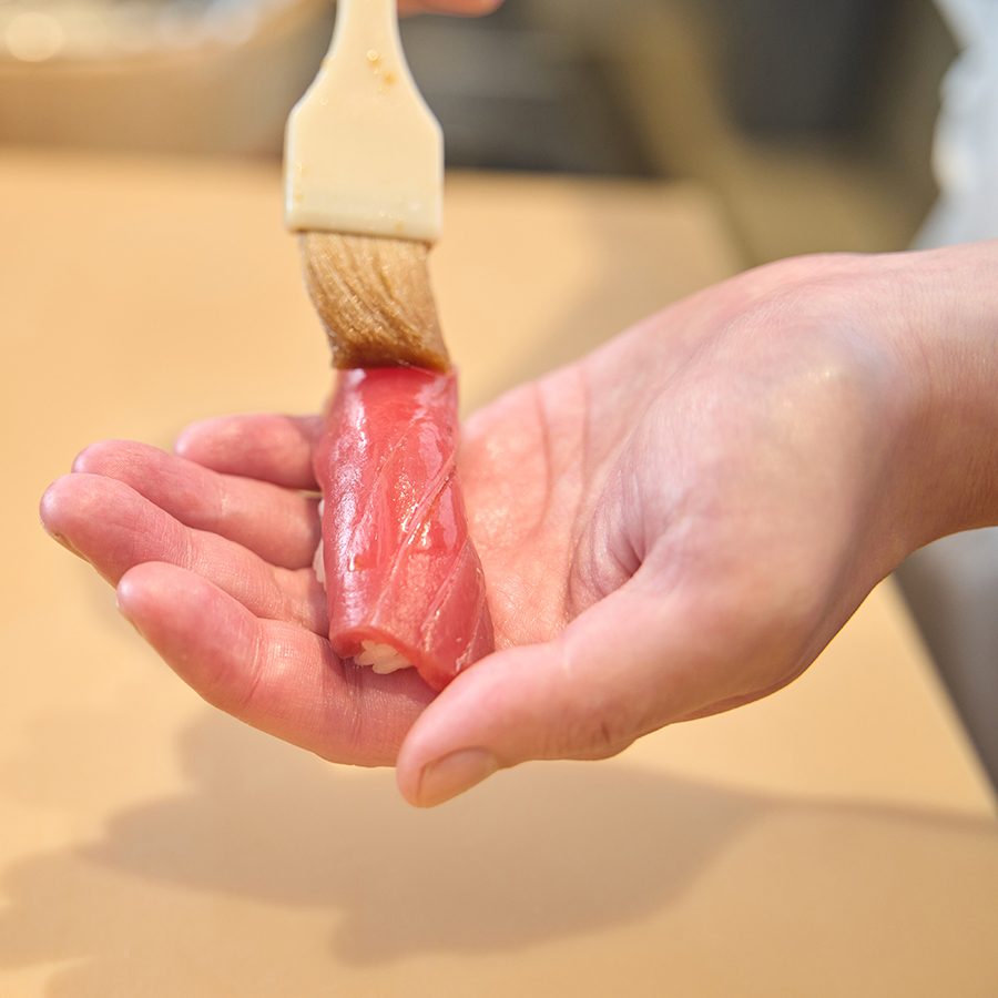 A hand holding a slice of sashimi with sauce being brushed onto it. 