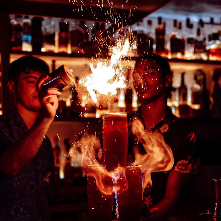 Three drinks are stacked on a bar with flames coming from them. Two men stand behind the bar. One is shaking powder onto the drinks, adding to the flames. 