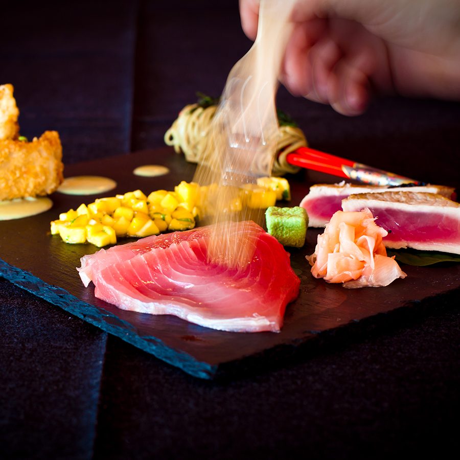 A hand hovering over a black stone slab plate with sashimi, soba, salad and tempura on it. 