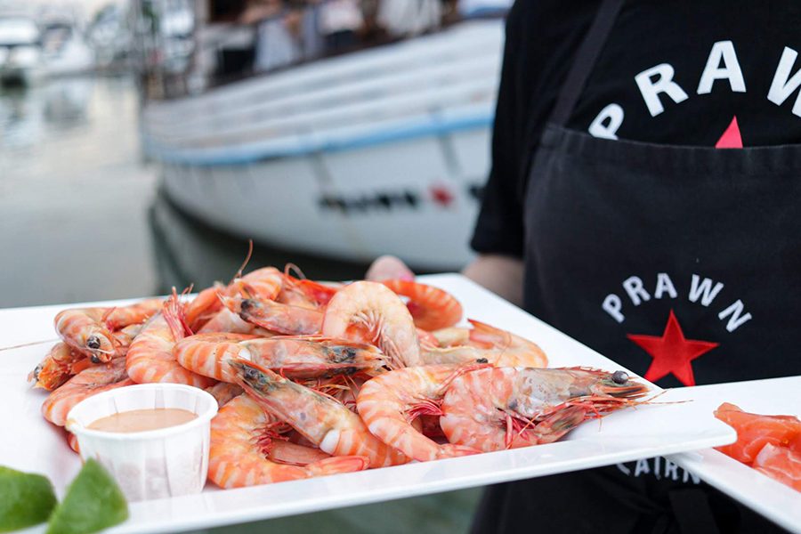A plate of shrimp with a small bowl of sauce. The person holding the plate is wearing a black Prawn Star T-shirt and apron.  