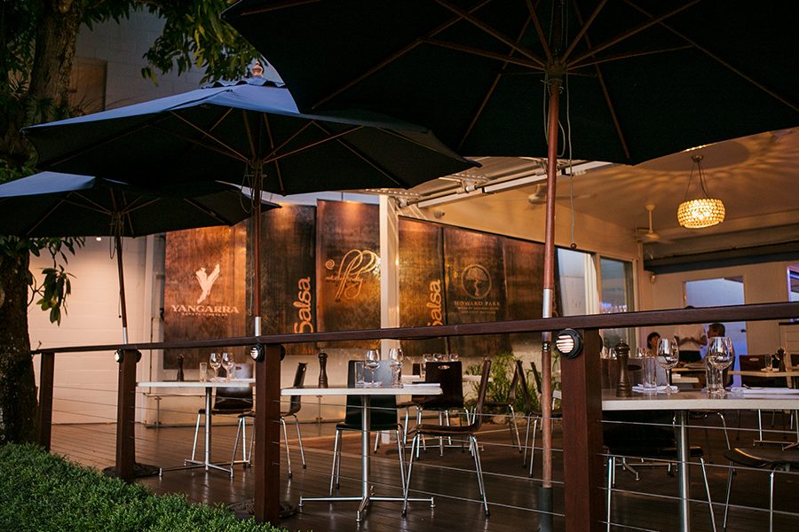 Tables and chairs and three large blue umbrellas in the outdoor dining area at Salsa Bar & Grill. Inside, a waiter is talking to a seated couple.  