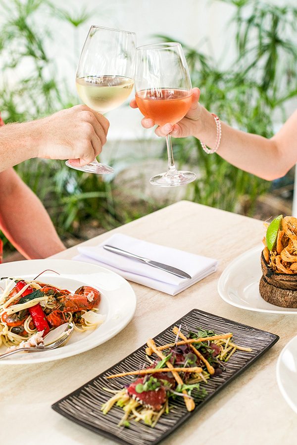 A close-up of two people clinking wine glasses over a table with three plates of food.