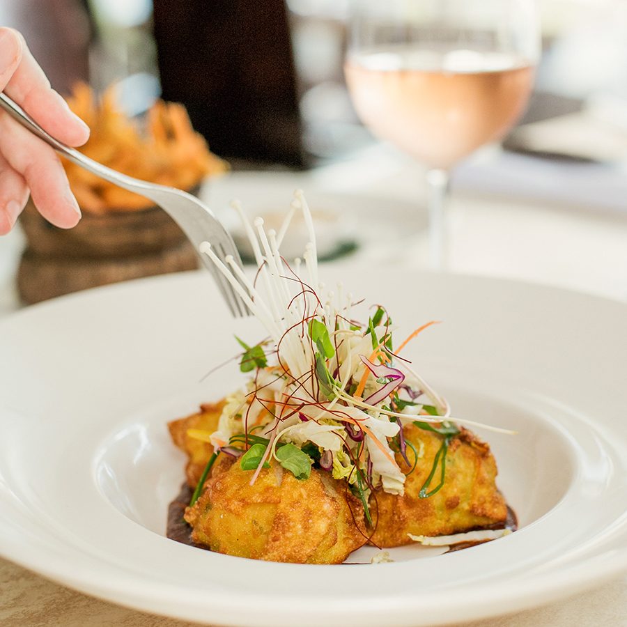 A hand holding a fork over a plate of food with a glass of wine in the background. 