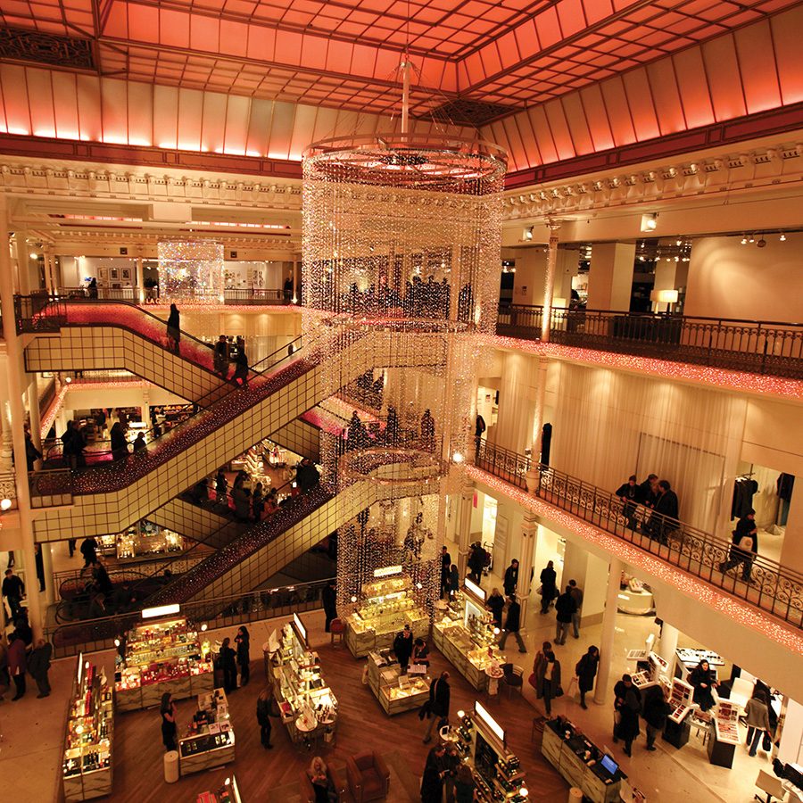 Interior image of Le Bon Marché, with a chandelier hanging from the  ceiling and escalators going up and down its floors