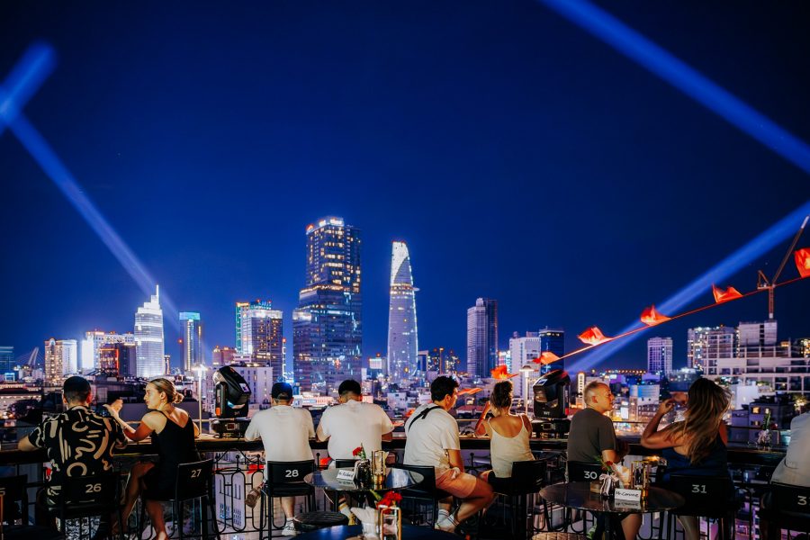A crowd of people enjoying a view of the skyline on the rooftop of Twilight Sky Bar Saigon in the evening with a light show display in the sky.