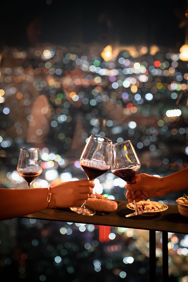 Two people clinking two glasses of red wine on the rooftop of Blank Sky Lounge at night with snacks and a view of Ho Chi Minh City’s evening skyline in the backdrop. 