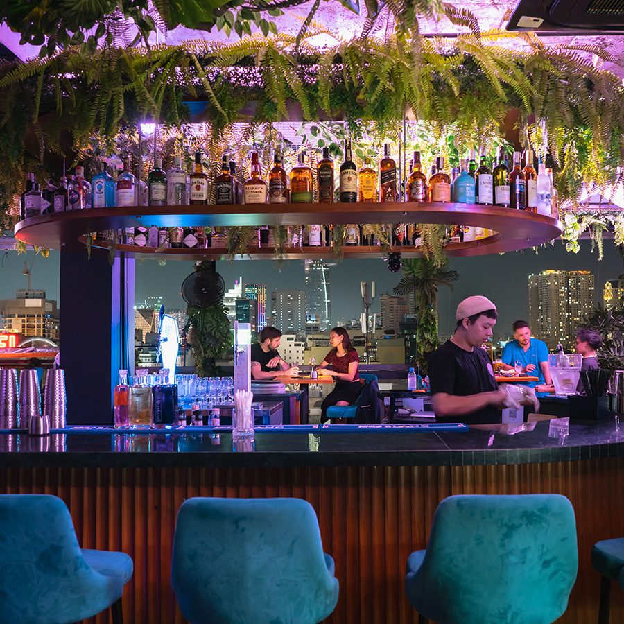 A bartender preparing drinks behind the open air bar at Cocoon Rooftop at night with a view of Ho Chi Minh City skyline in the backdrop. 
