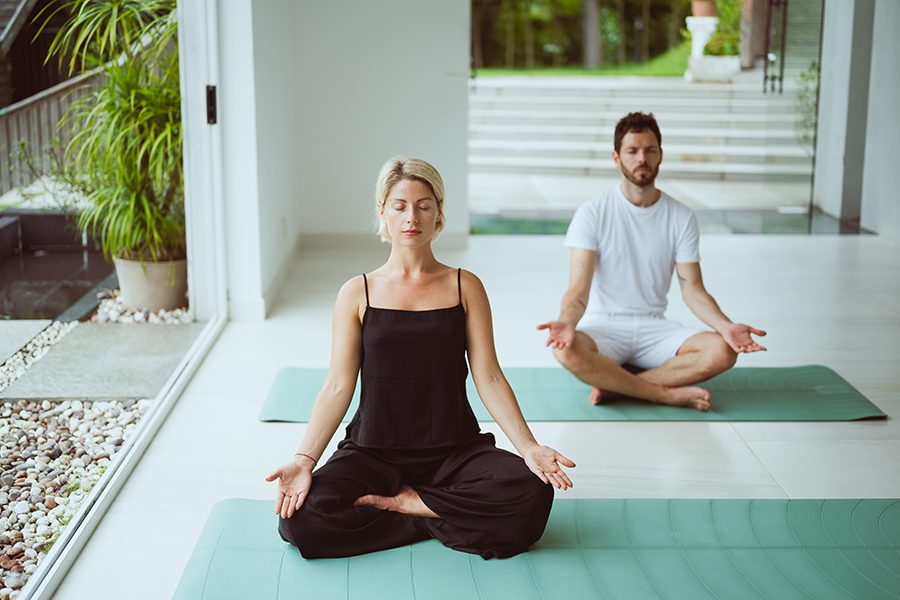 A woman dressed in black and a man dressed in white sit cross-legged with their eyes closed on green yoga mats.