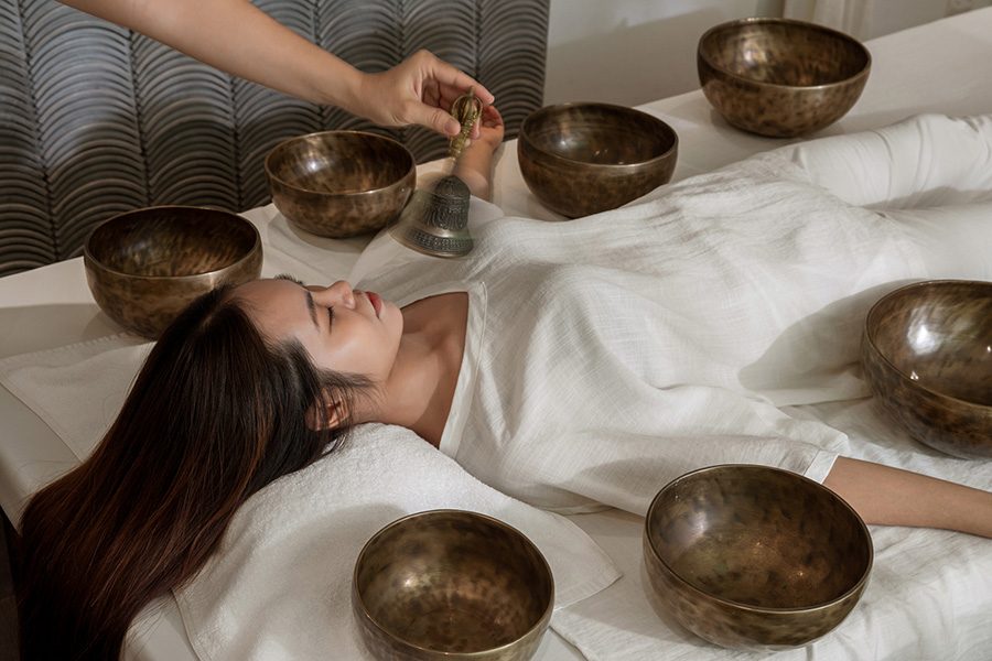 A woman lying down with bowls around her body while an outreached hand waves a bell over her.  
