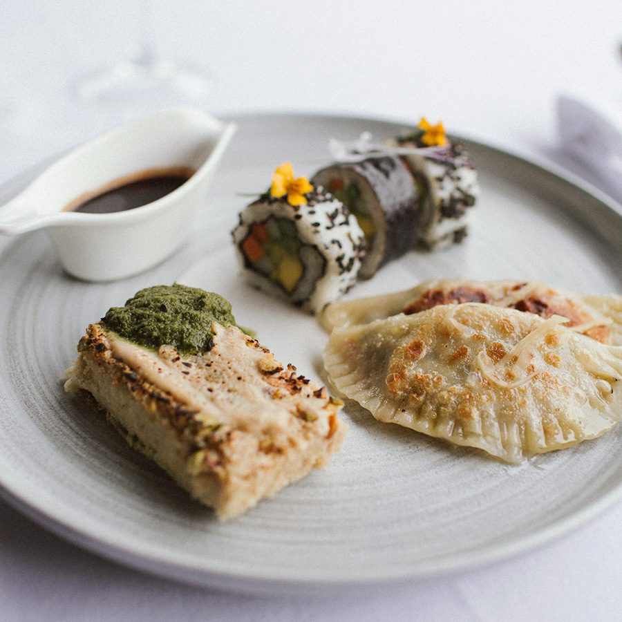 Food such as sushi and dumplings on a white plate on a white table, with a glass behind it. 