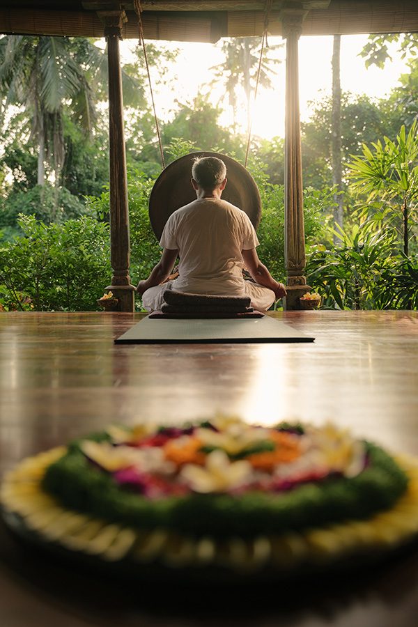 A man sitting cross-legged on a mat in front of a gong, facing the greenery outside.