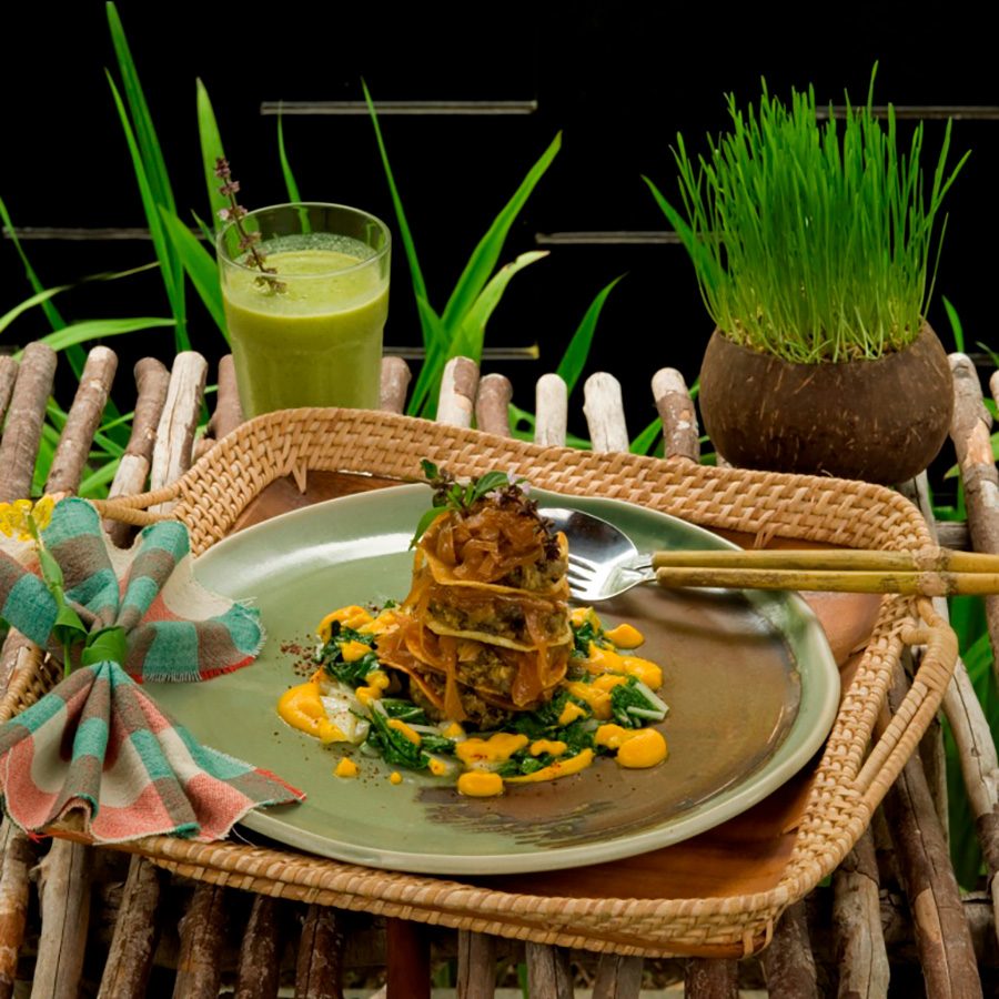 Food and utensils on a green plate on a woven tray. Behind it, there is a green smoothie and a small plant. 