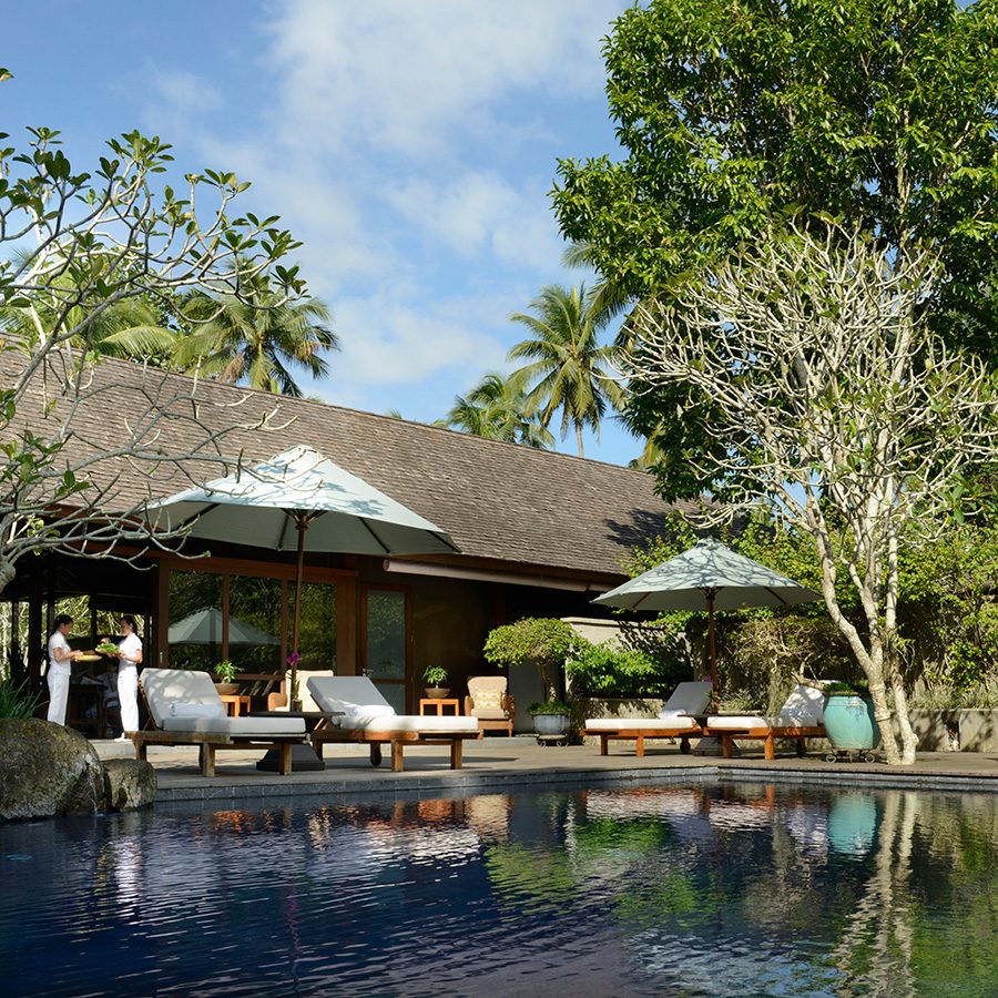 A pool lined with lounge chairs, patio umbrellas and trees. 