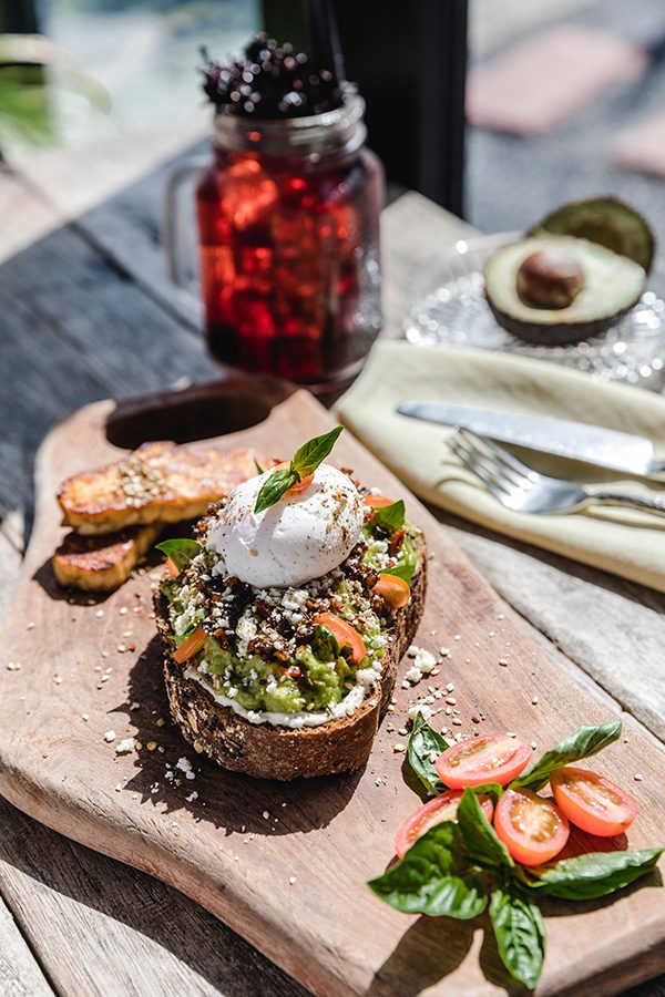 Avocado toast on a wooden serving board, with utensils, a drink and an avocado placed behind it. 