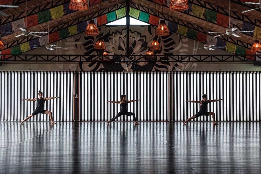 Three people dressed in black doing a Warrior II yoga pose in a room with a colourful patterned ceiling. 