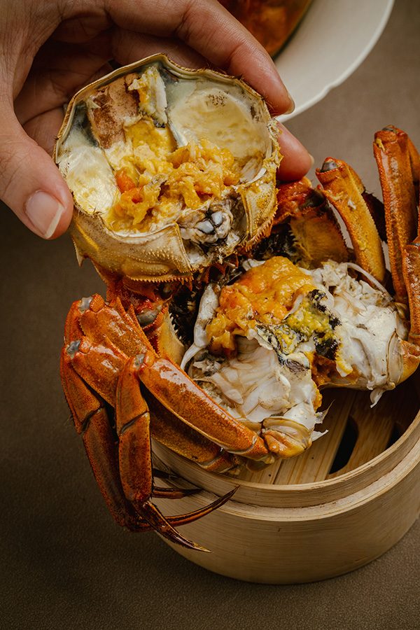 A closeup of a person holding the top shell of a hairy crab, displaying the crab meat, over a bamboo steamer basket, served at Chinese restaurant Moments Together in Hong Kong 