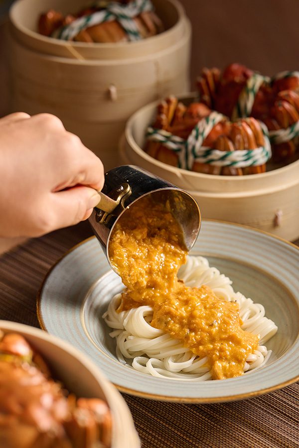 A closeup of a person pouring a small saucer of shrimp and hairy crab roe on top of a plate of braised inaniwa udon noodles with steamed hairy crabs in the background, featured on the set menu at Tze Yuet Heen in Tsuen Kwan O, Hong Kong 