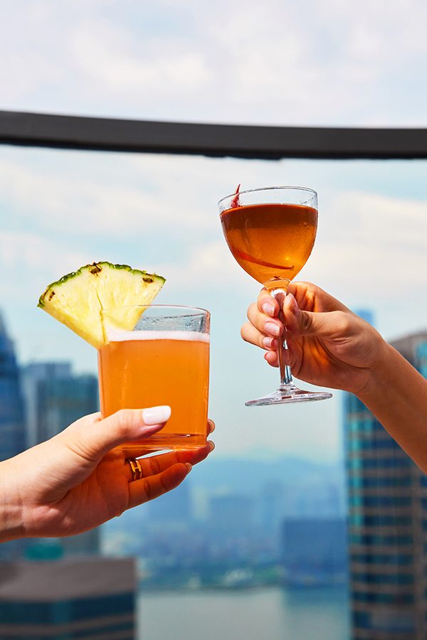 Close-up of two women’s hands holding an orange-coloured cocktail each. 