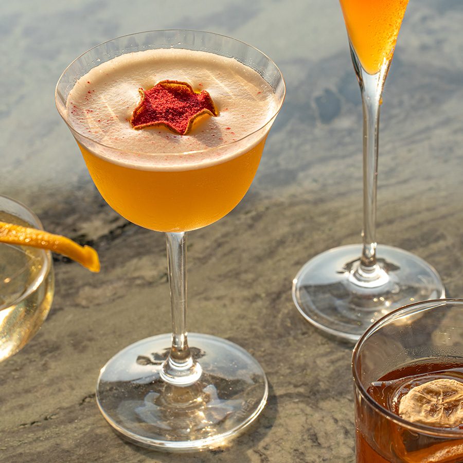 Four cocktails with yellow to orange colouring on a marble tabletop.