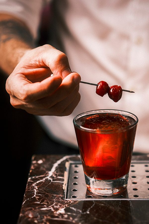 Close-up of a man holding a cherry garnish over a red-coloured cocktail. 