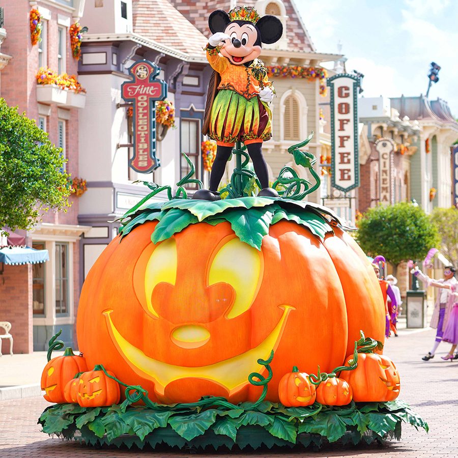 Mickey Mouse dressed in a pumpkin outfit stands on top of a pumpkin-themed float.