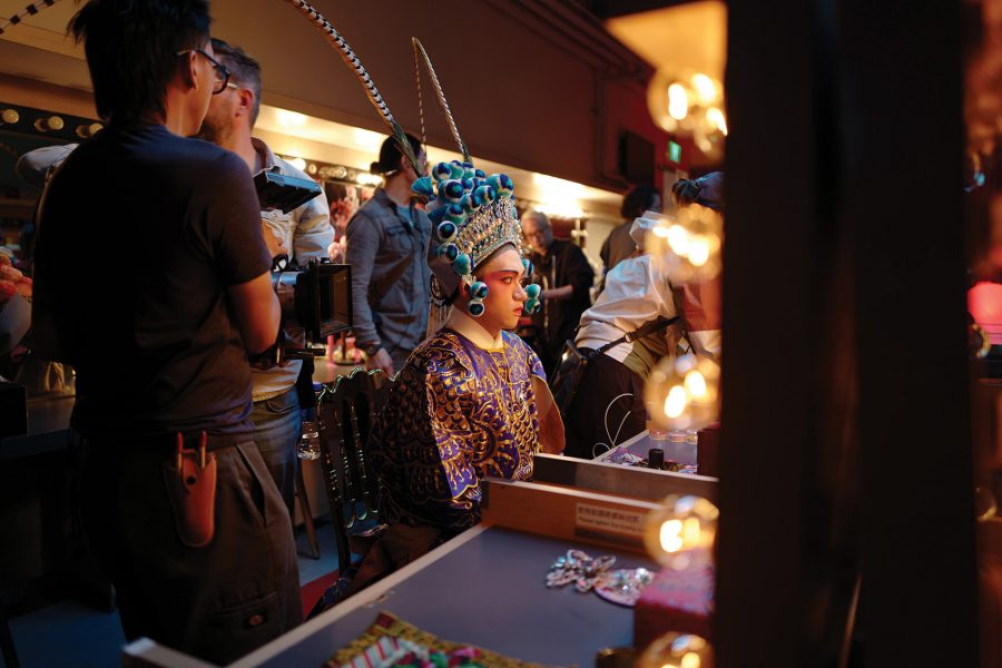 A Cantonese opera singer in costume sitting at his dressing table, while the film crew walks around on set for the Cathay Pacific's safety video. 