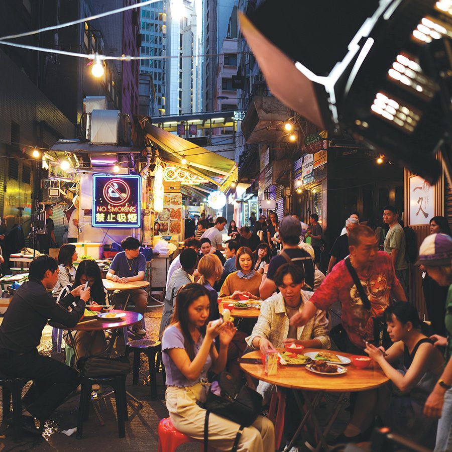 Multiple tables of people eating outside at a dai pai dong, with a neon “No Smoking” sign in the background.