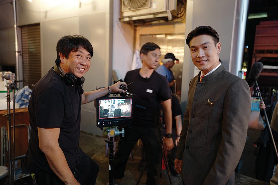 Two men smiling while on set. The man on the left is wearing headphones and holding onto a camera monitor, while the man on the right is Cathay Pacific cabin crew member Terry Lee, who is dressed in a grey uniform.  