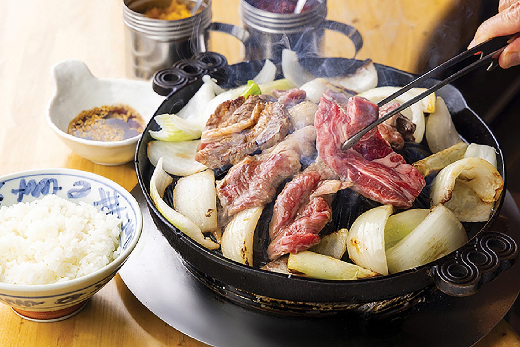 A  close-up of a person holding a pair of chopsticks and grilling meat and sliced onions on a hot plate on a dining table served with a bowl of white rice and dipping sauce served at Daruma Honten