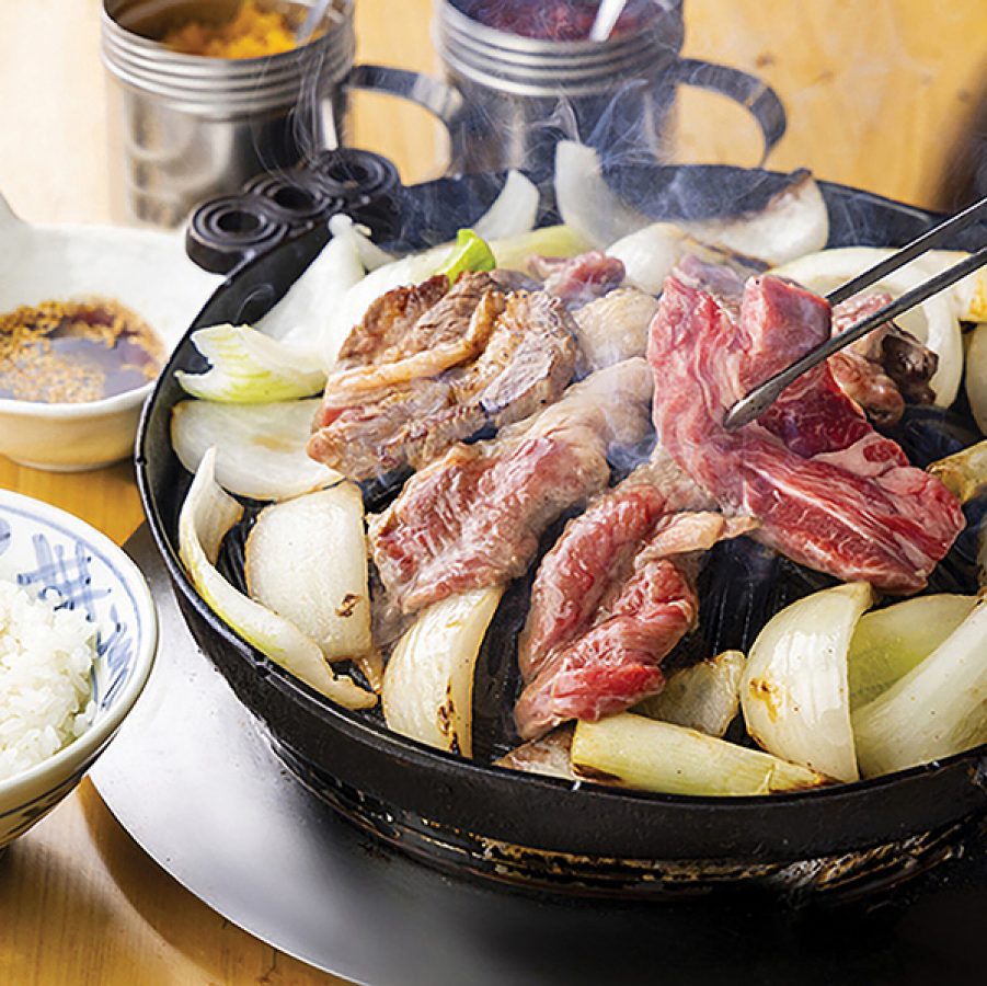 A  close-up of a person holding a pair of chopsticks and grilling meat and sliced onions on a hot plate on a dining table served with a bowl of white rice and dipping sauce served at Daruma Honten