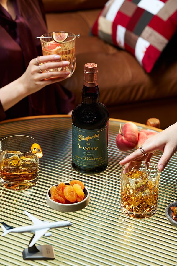 A bottle of Glenfarclas for Cathay Highland Single Malt Scotch is on a table along with glasses of whisky, a model airplane and a dish of apricots. A woman holds a glass of whisky on the left of the frame; another woman reaches for a glass on the right of the frame.