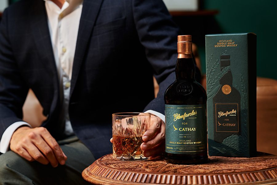 A man in a suit holds a glass of Glenfarclas for Cathay Highland Single Malt Scotch. The bottle and box are next to the glass on the wooden table. 